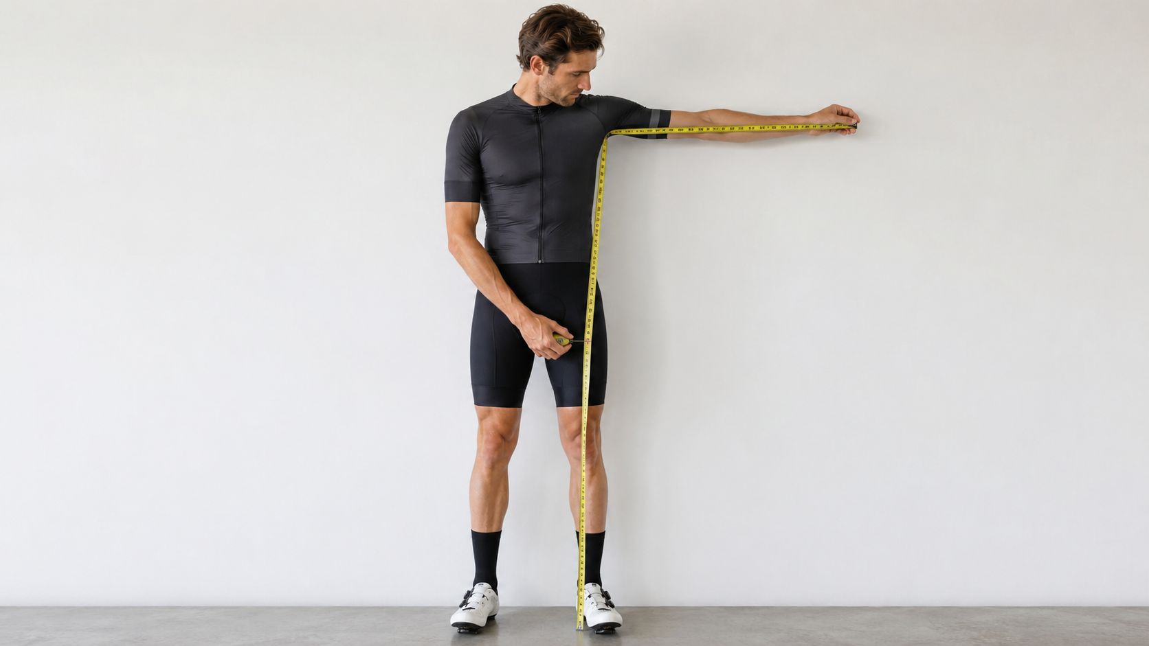 A professional male cyclist measuring his reach and leg length with a yellow measuring tape indoors.